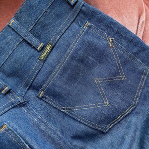 May include: Close-up of dark blue denim jeans. The back pocket features a distinctive 'W' stitch design in a contrasting yellow thread. The Wrangler logo is visible on the back waistband. The jeans are new and unworn.
