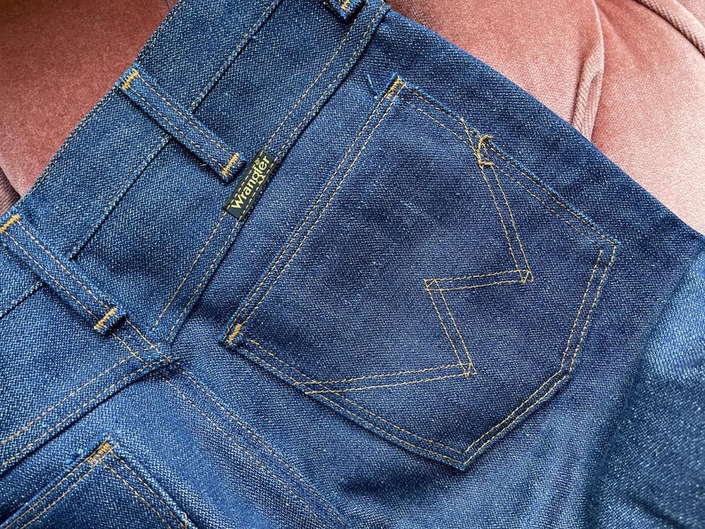 May include: Close-up of dark blue denim jeans. The back pocket features a distinctive 'W' stitch design in a contrasting yellow thread. The Wrangler logo is visible on the back waistband. The jeans are new and unworn.