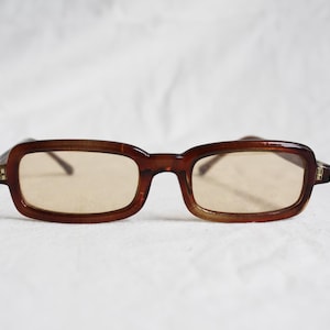 May include: Brown rectangular eyeglasses with light brown lenses. The frames have a subtle, rounded rectangular shape. The glasses are set against a white background, highlighting their vintage style.