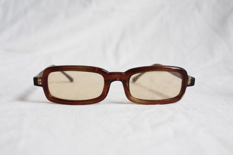 May include: Brown rectangular eyeglasses with light brown lenses. The frames have a subtle, rounded rectangular shape. The glasses are set against a white background, highlighting their vintage style.
