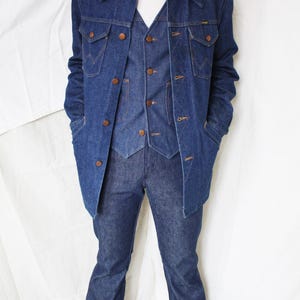 May include: A denim three-piece suit, including a jacket, vest, and flared jeans. The jacket has large pockets and brown buttons. The vest has a V-neck and matching buttons. The jeans are a dark wash denim.