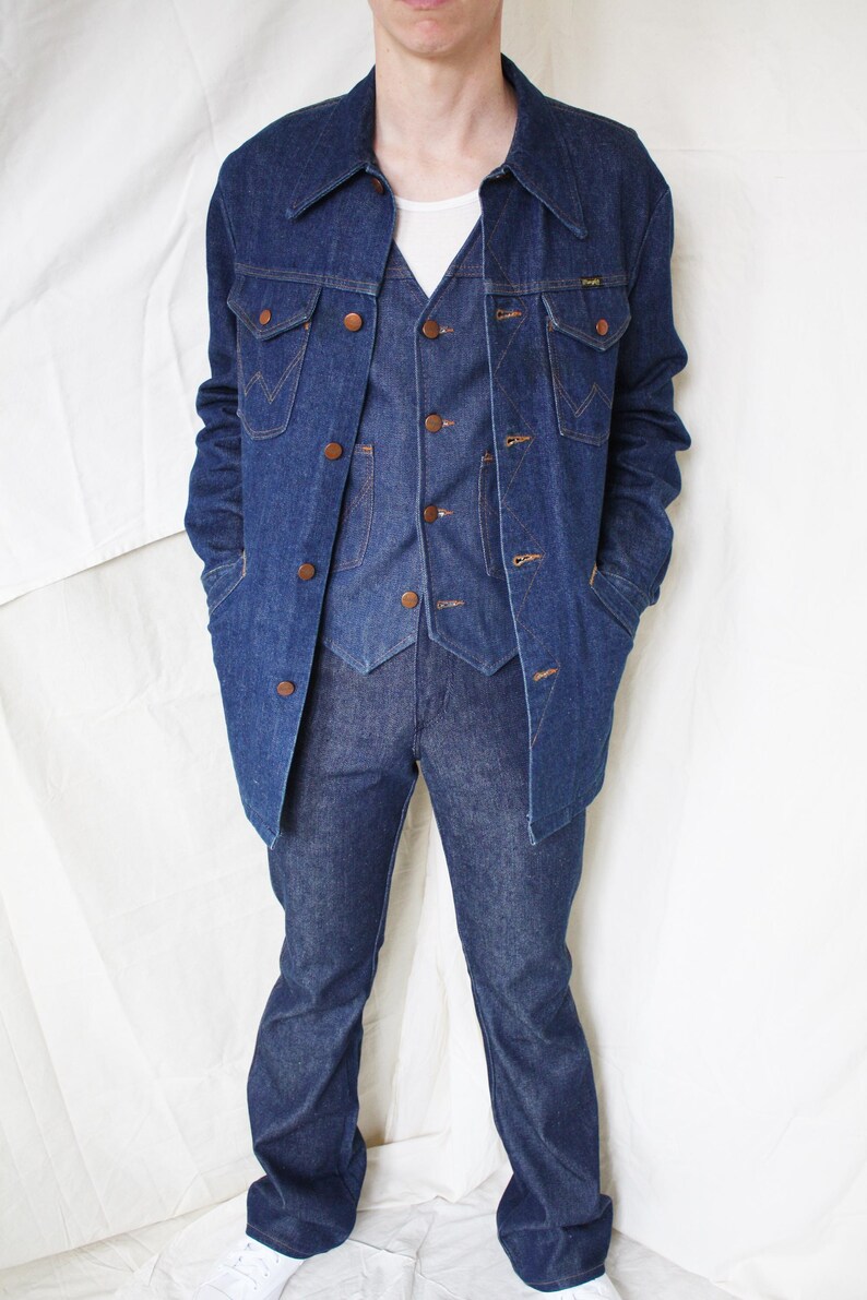 May include: A denim three-piece suit, including a jacket, vest, and flared jeans. The jacket has large pockets and brown buttons. The vest has a V-neck and matching buttons. The jeans are a dark wash denim.