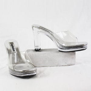 May include: Clear platform heels with a silver sole and a transparent strap. The heels have a chunky, clear block heel. The word "temptation" is printed on the insole. The shoes are displayed on a small, grey rectangular block.