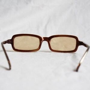 May include: Brown rectangular eyeglasses with light-tinted lenses. The frames are a dark brown color, and the temples are a lighter brown with silver-colored arms. The glasses are set against a white background.
