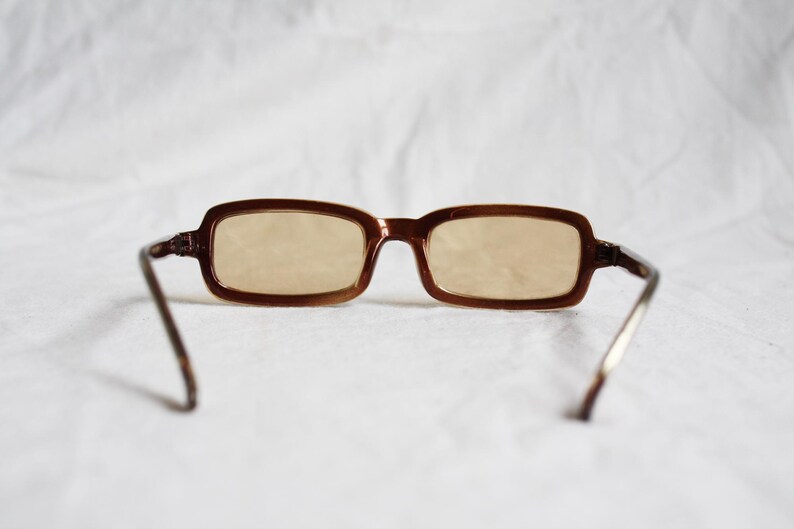 May include: Brown rectangular eyeglasses with light-tinted lenses. The frames are a dark brown color, and the temples are a lighter brown with silver-colored arms. The glasses are set against a white background.