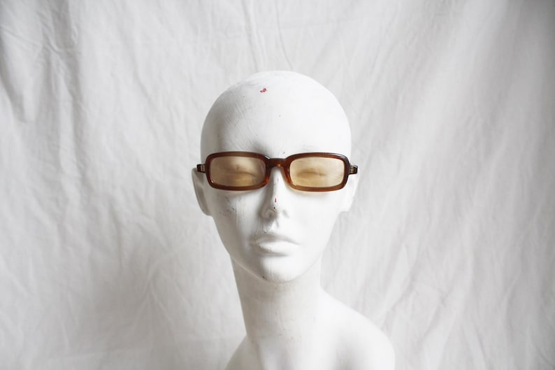 May include: Brown rectangular sunglasses with amber lenses are displayed on a white mannequin head. The glasses have a classic, retro design with a dark brown frame. The background is a plain white cloth.