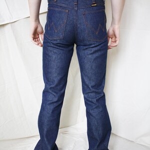 May include: Dark blue denim jeans with orange stitching and a Wrangler logo on the back pocket. The jeans have a classic bootcut style. The person is wearing a white tank top.