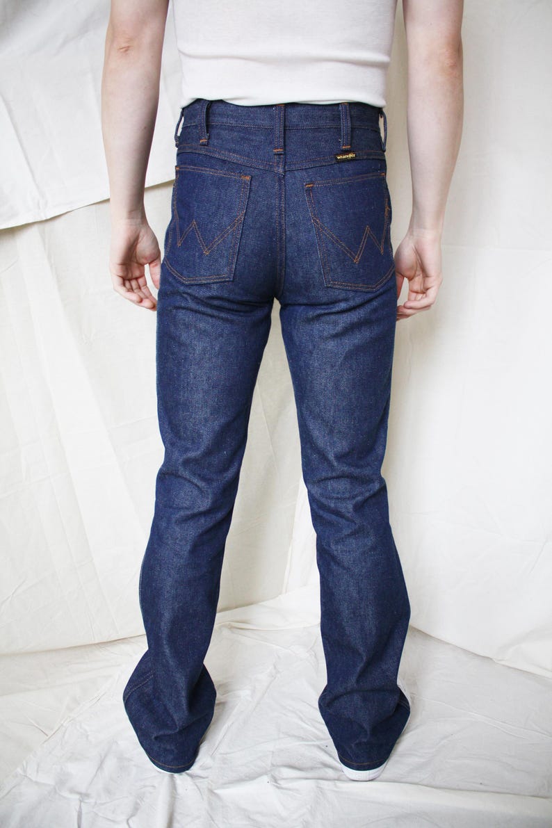 May include: Dark blue denim jeans with orange stitching and a Wrangler logo on the back pocket. The jeans have a classic bootcut style. The person is wearing a white tank top.