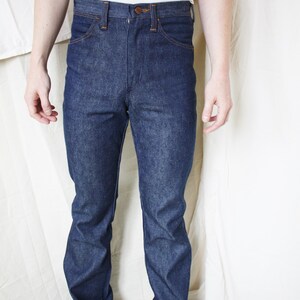 May include: Dark blue denim jeans with a classic five-pocket design. The jeans have a button and zipper closure, and a straight leg fit. The model is wearing a white t-shirt and white sneakers.
