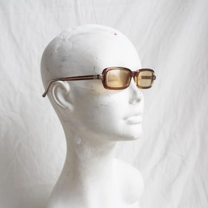 May include: Brown rectangular sunglasses with tinted lenses are displayed on a white mannequin head. The frames have a dark brown color with a lighter brown accent on the temples. The lenses have a yellow tint.