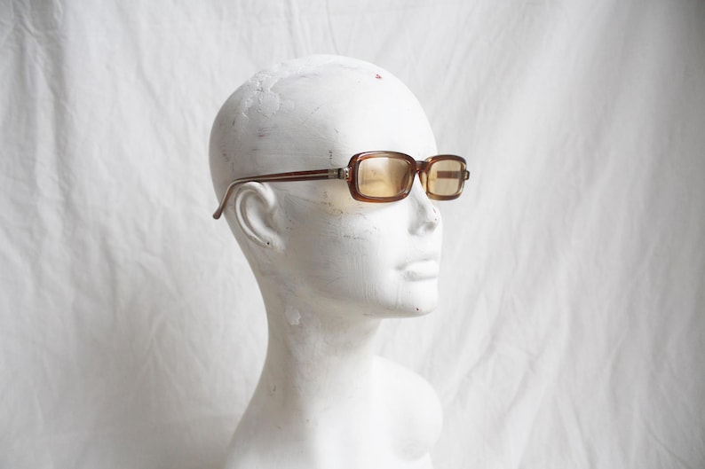 May include: Brown rectangular sunglasses with tinted lenses are displayed on a white mannequin head. The frames have a dark brown color with a lighter brown accent on the temples. The lenses have a yellow tint.