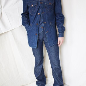May include: A denim three-piece suit, including a jacket, vest, and flared-leg pants. The jacket has two chest pockets and button closures. The suit is a dark blue wash, and the model is wearing white sneakers.