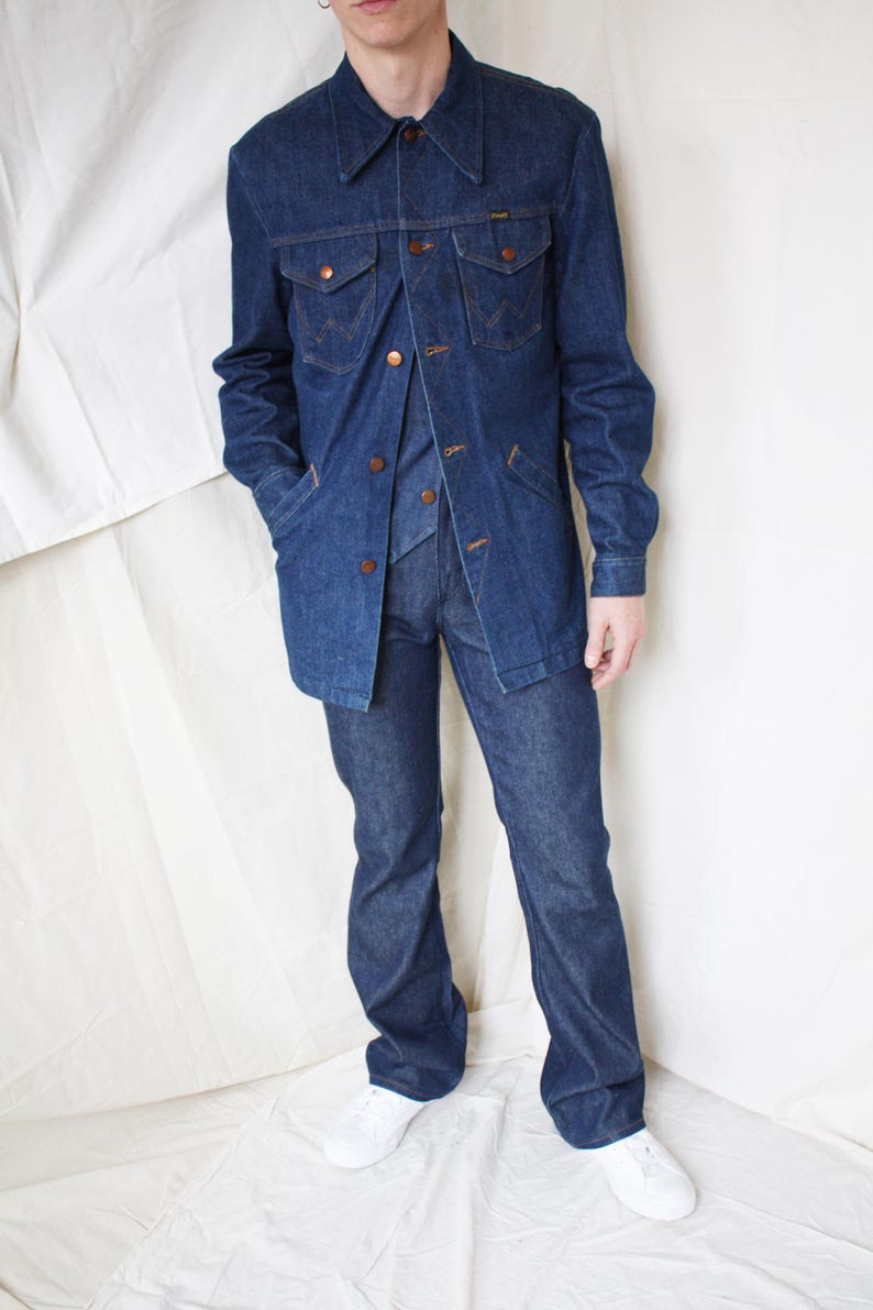 May include: A denim three-piece suit, including a jacket, vest, and flared-leg pants. The jacket has two chest pockets and button closures. The suit is a dark blue wash, and the model is wearing white sneakers.