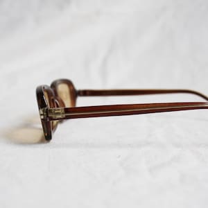 May include: Brown eyeglasses with a rectangular frame and thin temples. The glasses are set against a white background, showcasing their classic design. The lenses are a light brown tint.