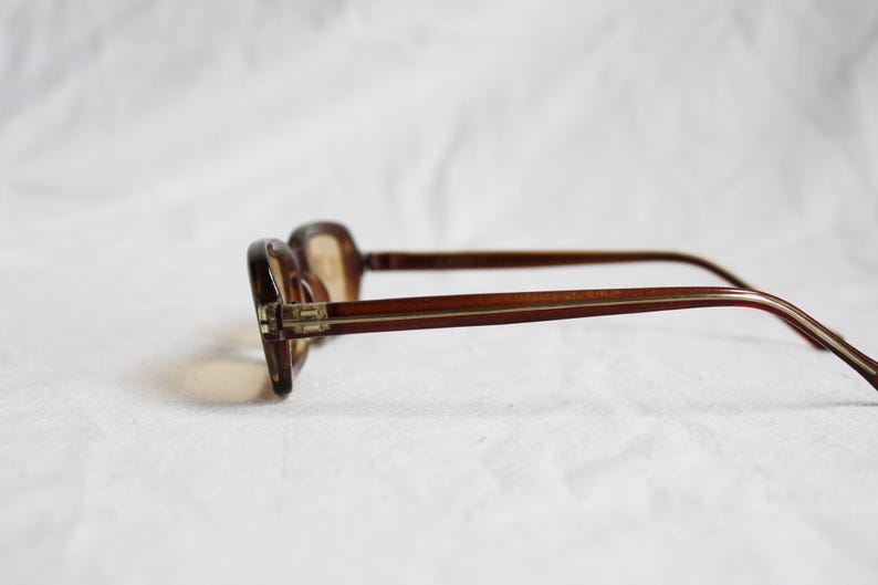 May include: Brown eyeglasses with a rectangular frame and thin temples. The glasses are set against a white background, showcasing their classic design. The lenses are a light brown tint.