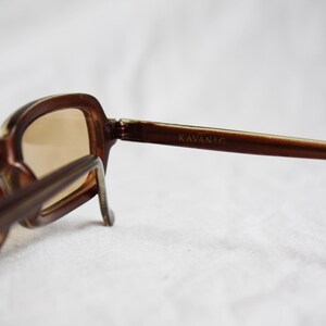 May include: Brown-framed sunglasses with tinted lenses. The sunglasses have a rectangular shape and the brand name "KAVANA" is visible on the temple. The glasses are set against a white background.