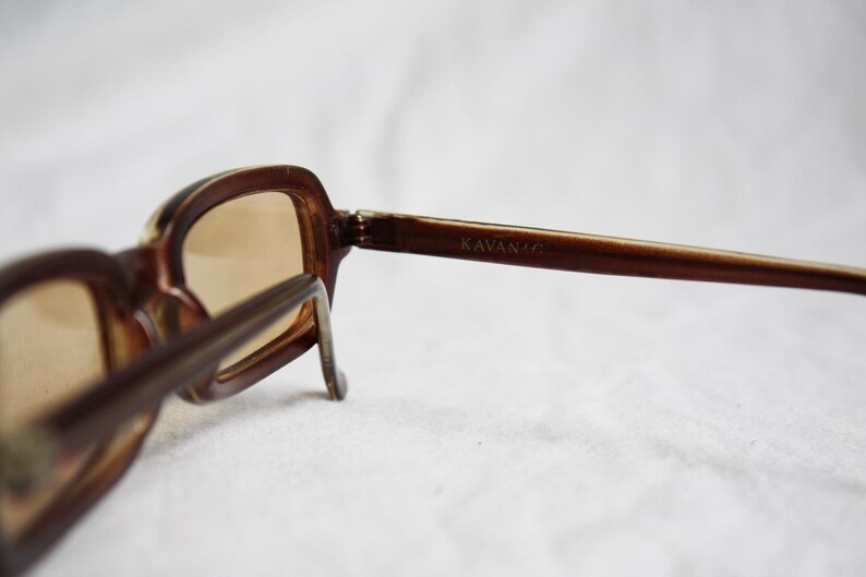 May include: Brown-framed sunglasses with tinted lenses. The sunglasses have a rectangular shape and the brand name "KAVANA" is visible on the temple. The glasses are set against a white background.