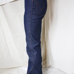 May include: Dark blue denim jeans with a classic five-pocket design. The jeans have a straight leg fit and are worn with white sneakers. The back pocket features a distinctive stitching pattern. The jeans are made of a sturdy denim material.