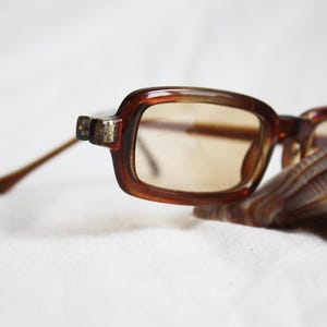May include: Brown rectangular eyeglasses with tinted lenses rest on a seashell. The glasses have a thick, dark brown frame and slender, light brown arms. The lenses are a light brown color.