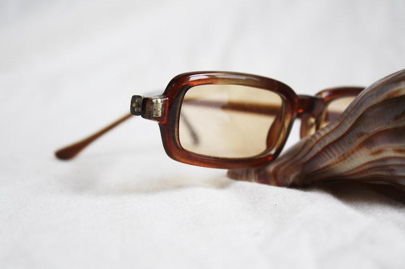 May include: Brown rectangular eyeglasses with tinted lenses rest on a seashell. The glasses have a thick, dark brown frame and slender, light brown arms. The lenses are a light brown color.