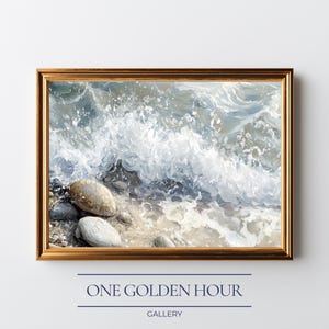 May include: A framed painting of a seascape with a golden frame. The artwork depicts a wave crashing on a sandy shore with pebbles. The color palette includes shades of blue, white, and beige. The text "ONE GOLDEN HOUR GALLERY" is at the bottom.