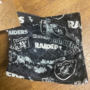 May include: A black and white Raiders-themed bowl cozy. The fabric features the Raiders logo and team name in a repeating pattern. The cozy is quilted with white thread and has a square shape.