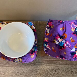 May include: Two purple fabric pot holders with a floral and cartoon character design. The pot holders feature a white bowl on top of one of them.