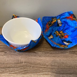 May include: A set of two blue and orange fabric bowl cozies with a print of cartoon monster trucks. The cozies are designed to keep bowls warm.