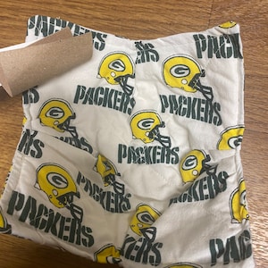 May include: A white fabric pot holder with a green and yellow pattern of the Green Bay Packers logo. The logo features a football helmet with a green "G" on the side and the word "Packers" written in black.