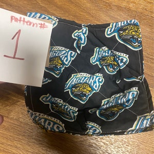 May include: Black quilted hot pad with a repeated pattern of the Jacksonville Jaguars logo. The logo is in teal, yellow, and white with the words "Jacksonville" and "Jaguars" printed on it. The text "Pattern # 1" is written on a white piece of paper.
