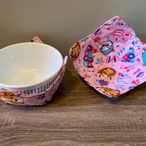 May include: Two pink fabric bowl cozies with a paw patrol print. The cozies are made of cotton fabric and have a quilted design.