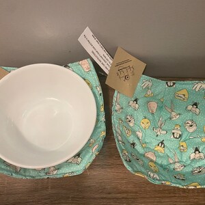 May include: Two turquoise blue fabric bowl cozies with a white background and a cartoon print of Bugs Bunny, Daffy Duck, Tweety Bird, Sylvester the Cat, Porky Pig, and other Looney Tunes characters. The cozies are made of quilted fabric and have a white stitching around the edges.