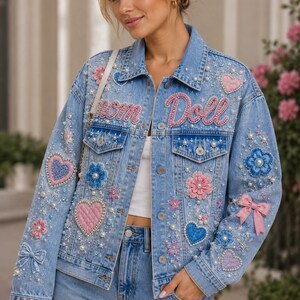 May include: A light blue denim jacket featuring the text "Mom Doll" in pink beaded lettering. The jacket is decorated with various embroidered patches including pink and blue hearts, flowers, and bows, along with scattered white pearl embellishments.