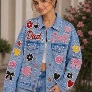 May include: A light blue denim jacket featuring the text Dad Doll in red and pink script. The jacket is heavily embellished with various colorful patches including hearts, flowers, stars, and bows, accented with scattered white faux pearls.