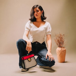 Può includere: Un top bianco a maniche corte con scollo a V, jeans blu scuro e una borsa in stile cartone animato con corpo grigio chiaro, patta superiore rosso brillante, chiusura gialla e spessi contorni neri che creano un aspetto grafico bidimensionale.