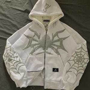 May include: White zip-up hoodie with a spiderweb design on the front and sleeves. The hoodie features a hood with a logo and a soft, light-coloured interior. The design is in a light grey colour.