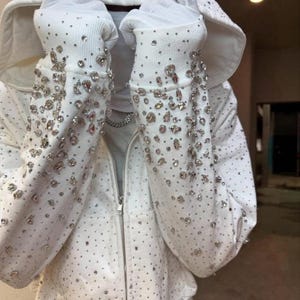 Puede incluir: Sudadera blanca con cremallera y pedrería dispersa. Las mangas y la capucha están adornadas con pedrería brillante más grande. La sudadera tiene una cremallera plateada y texto en la capucha.