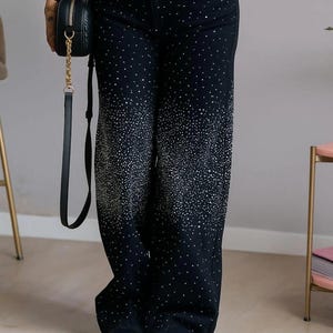 Puede incluir: Vaqueros negros de pernera ancha con un patrón brillante y punteado que se desvanece hacia la parte inferior. Los vaqueros se combinan con zapatos negros y un pequeño bolso negro con una correa de cadena dorada. El estilo general es informal y moderno.