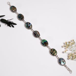 Könnte beinhalten: Ein silberfarbenes Gliederarmband mit sieben ovalen Abalone-Muschel-Cabochons. Jede irisierende Muschel zeigt wirbelnde Muster in Grün-, Blau- und Violetttönen, die in einer gewellten Metallfassung sitzen. Das Armband hat einen Karabinerverschluss.