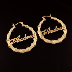 May include: A pair of gold-colored hoop earrings. Each earring features the name "Andrea" in a cursive font. The hoops have a bamboo-like design, with a clasp closure. The background is black.