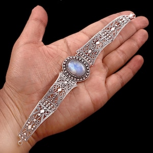 Puede incluir: Pulsera de plata ornamentada con un centro de piedra lunar ovalada e iridiscente. La pulsera tiene intrincados patrones de filigrana y puntos, con un diseño calado y delicado. La pulsera se muestra sobre un fondo oscuro.