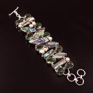 Può includere: Un bracciale in argento con una varietà di pietre preziose. Incorpora conchiglia di abalone, pietre verdi e piccole perle bianche. Il design del bracciale è ambientato su uno sfondo nero, evidenziando i colori e le trame dei gioielli.