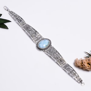 Könnte beinhalten: Ein silbernes Armband mit einem ovalen, hellblauen Stein in der Mitte. Das Armband besteht aus komplizierten, durchbrochenen Metallgliedern mit einer dunklen, oxidierten Oberfläche und wird mit einem kleinen Karabinerverschluss geschlossen.