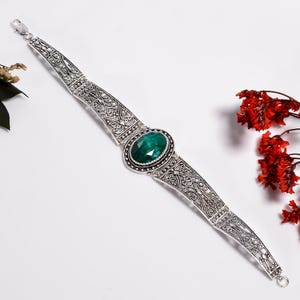 Könnte beinhalten: Ein silberfarbenes Armband mit einem ovalen, facettierten dunkelgrünen Stein in der Mitte. Das Armband besteht aus beweglichen Metallsegmenten mit komplizierten, durchbrochenen Filigranmustern und einem sicheren Karabinerverschluss.