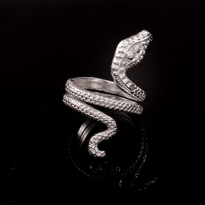 Op de afbeelding: Een zilverkleurige ring in de vorm van een slang, opgerold om de vinger. De kop van de slang is omhoog en het lichaam is getextureerd met schubben. De ring staat tegen een zwarte achtergrond.