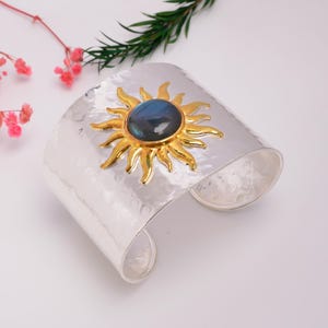 May include: A wide, hammered silver cuff bracelet featuring a central gold-toned sun motif. The sun design has radiating rays and holds a smooth, oval-shaped, dark blue iridescent gemstone at its centre, creating a bold and reflective appearance.