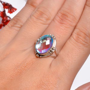 Può includere: Un anello in argento con una grande pietra ovale sfaccettata e iridescente che mostra uno spettro di colori tra cui blu, viola e rosa. La montatura è realizzata in argento lucido con piccole perline metalliche decorative attorno alla pietra.