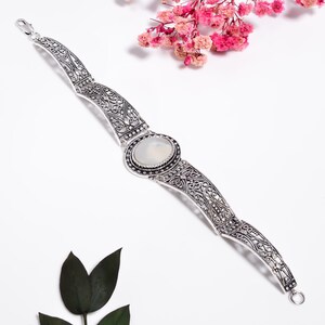 May include: A silver-toned metal bracelet featuring an oval-shaped, milky white cabochon stone at the center. The bracelet is composed of articulated, curved links with intricate, filigree-style cut-out patterns and a lobster claw clasp closure.