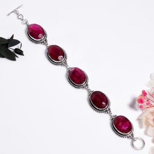 Könnte beinhalten: Ein Silberarmband mit fünf ovalen, tief magentafarbenen facettierten Edelsteinen. Jeder Stein ist in eine dekorative Silberfassung mit Perlenrand eingefasst. Das Armband wird mit einem silbernen Knebelverschluss und einem Ring geschlossen.