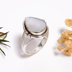 Puede incluir: Un anillo de plata de ley con una piedra preciosa de nácar blanco iridiscente en forma de pera, engastada en un bisel liso. La banda se divide en dos secciones cerca del engaste, decorado con pequeñas esferas de plata. El interior lleva el sello 925.